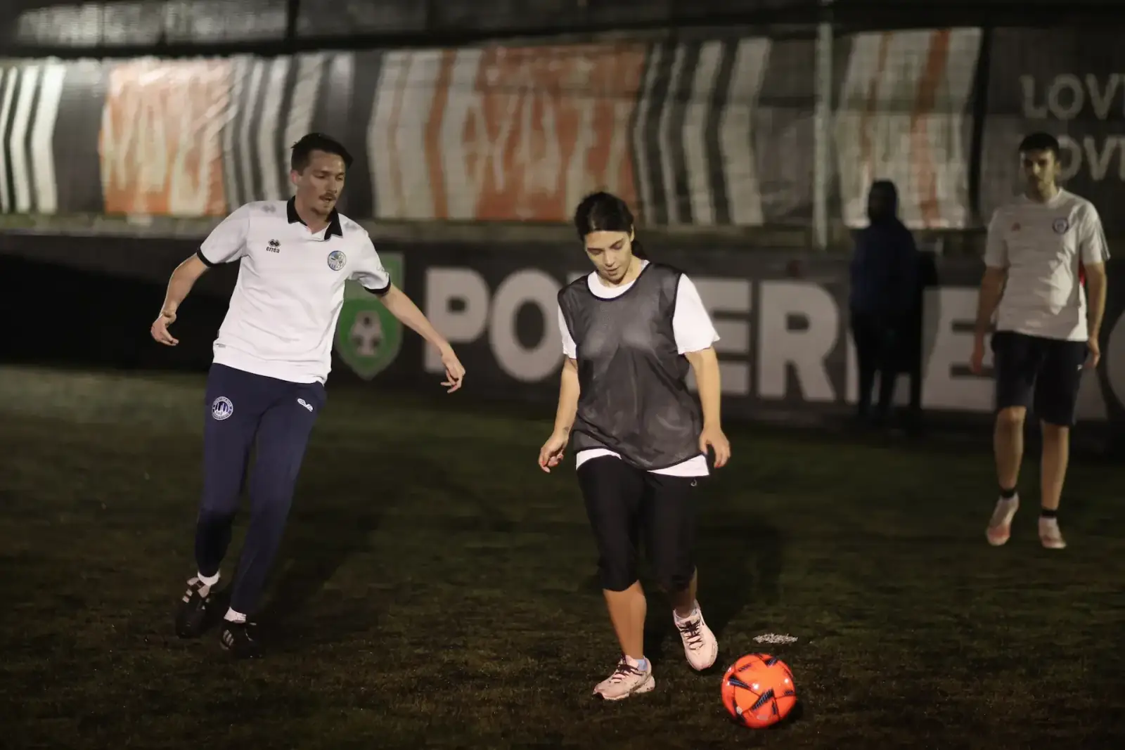 Mixed-team players challenging for the ball under floodlights on a five-a-side pitch, Powerleague advertising boards behind them.