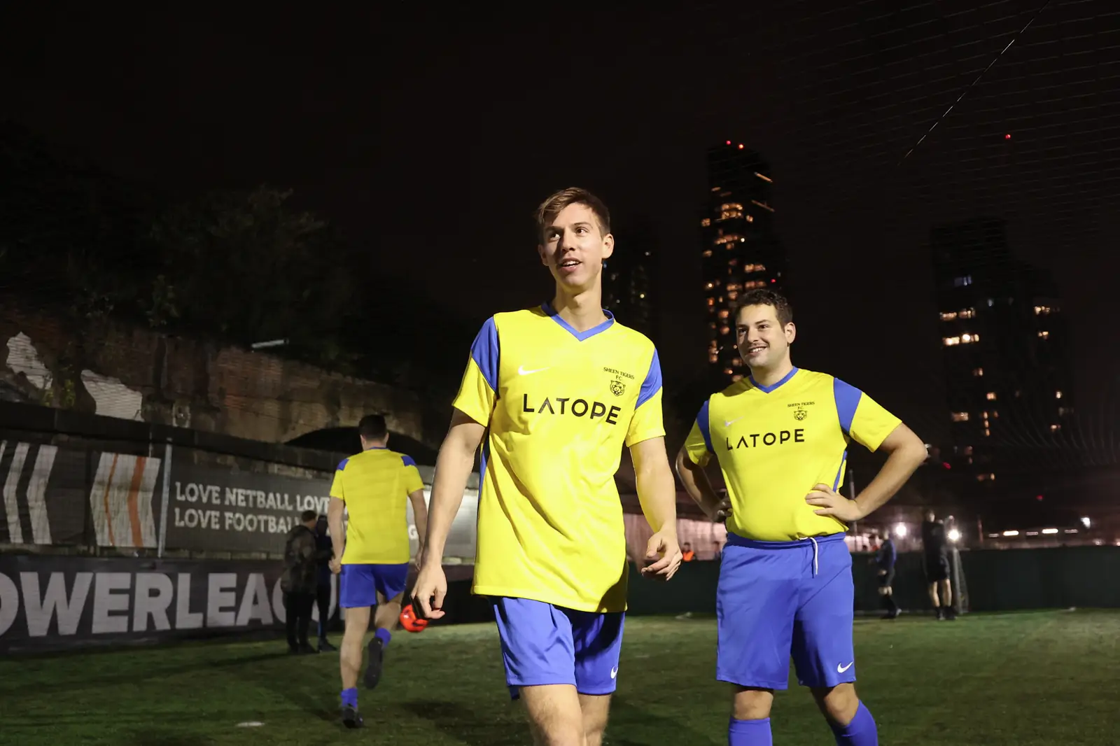 Two teammates in yellow and blue kit walking off a floodlit five-a-side pitch together, London tower blocks lit up behind them.