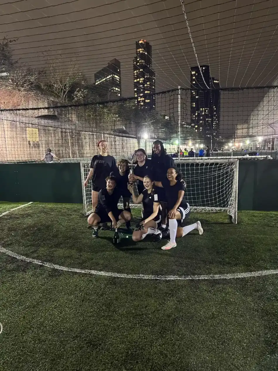 A women's five-a-side team kneeling and celebrating around a trophy in front of the goal, London tower blocks lit up at dusk behind them.